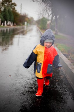 Yağmurda tek başına koşan küçük bir çocuk kalyuzhla koşar. Mavi, kırmızı ve turuncu renkli ve kırmızı çizmeli bir yağmurlukla. Mutlu çocuk. Tasasız çocukluk. Kötü hava. Yağmuru severim. sis.