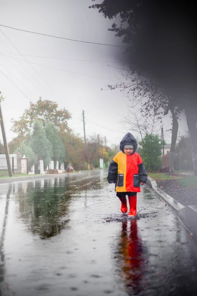 A small child, alone in the rain, runs on kalyuzh.In a raincoat in blue, red and orange colors and red boots. Happy child. Carefree childhood. Bad weather. I like rain. fog