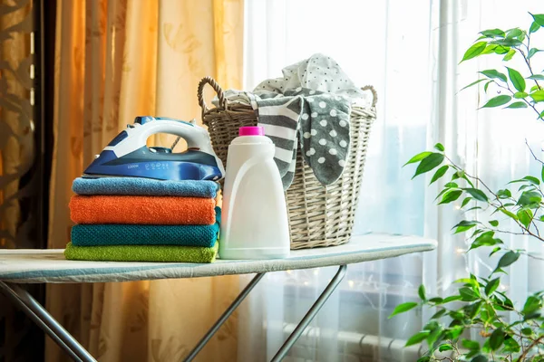 Electric iron and basket of clean linen on ironing board, against the window