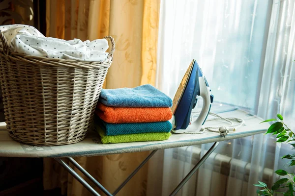 Electric iron and basket of clean linen on ironing board, against the window