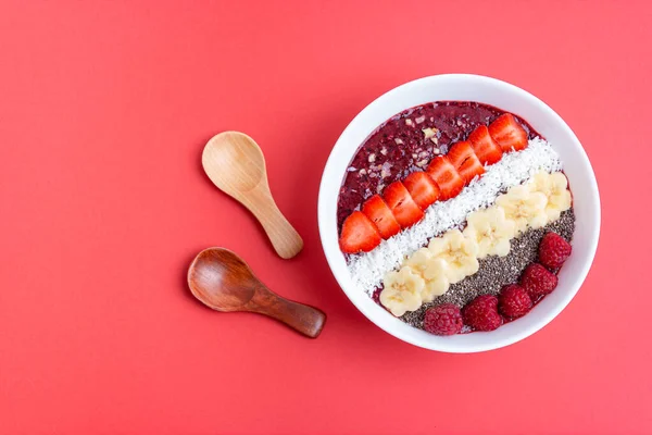 Smoothie bowl or acai berry bowl with chia, strawberries, raspberries, banana slices and coconut flakes. Healthy vegan food. Top view, red background.