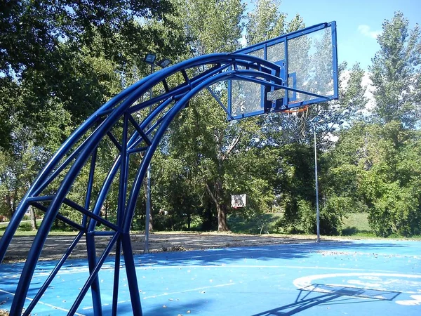 アスリートや人がいないバスケットボールのスポーツフィールド。バスケットとベンチ付きのバスケットボールタワーは青色に塗られています。夏、緑の葉を持つ木々. — ストック写真