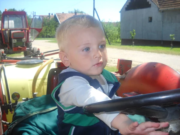 Un niño pequeño se sienta en el tractor de su padre. Un niño juega una volición de transporte. Primavera en el pueblo —  Fotos de Stock