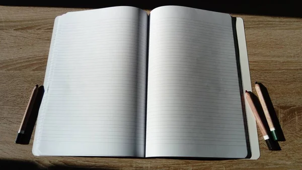 U-Γυρίστε το σημειωματάριο με κενές λευκές σελίδες. Στα αριστερά και δεξιά του σημειωματαρίου στο ξύλινο τραπέζι είναι χρωματιστά μολύβια. — Φωτογραφία Αρχείου
