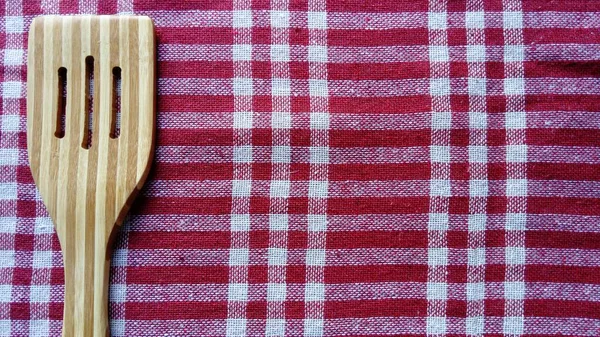 A wooden spatula for stirring cooked food lies on a cotton tablecloth. The kitchen tablecloth is made of red-white checked fabric. Fabric tartan — Stock Photo, Image