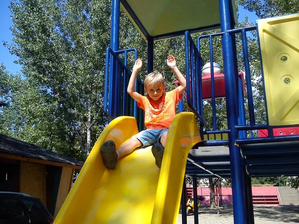 A boy got ready to roll off the plastic slide in the playground. The boy raised his hands and smiles.