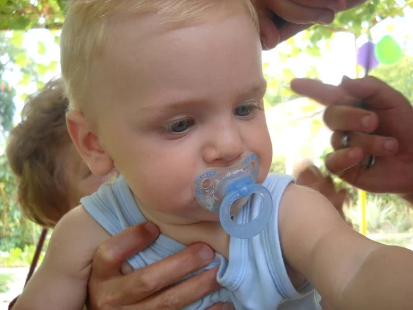 Крупним планом малюки голову білої раси з блакитними очима з соску в роті. Дитяче волосся обробляють ножицями. Руки перукаря і бабусі — стокове фото
