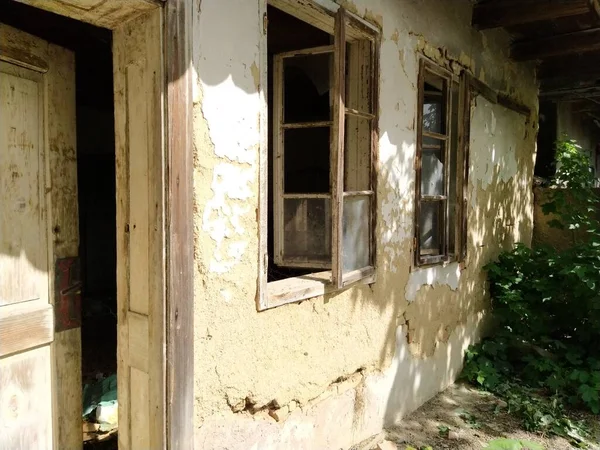 Τοίχος με παλιά παράθυρα και μια πόρτα σε ένα ετοιμόρροπο αγροτόσπιτο. Η έννοια της αστικοποίησης και της λήθης της πατρίδας. Εγκαταλελειμμένο σπίτι. Σπασμένο γυαλί στα παράθυρα και γύψος που πέφτει — Φωτογραφία Αρχείου
