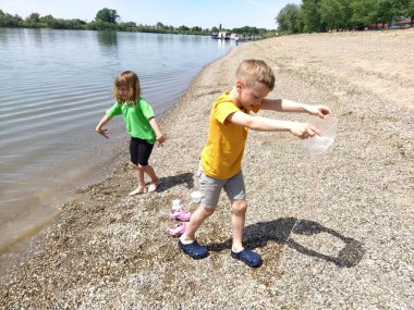 Sremska Mitrovica, Sırbistan, 30 Mayıs 2020, plajda bir oğlan ve bir kız oynuyorlar. Çocuklar nehir kıyısında su, kum ve çakıl taşlarıyla oynarlar. Çocuklar sade tişört giyer. Sırbistan, Sava nehri. Sarı saçlı çocuklar.