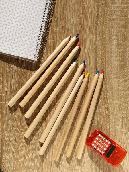 Vista dall'alto di quaderno bianco, calcolatrice e matite su sfondo di legno. Cancelleria d'ufficio. Materiale scolastico per lo studente. Concetto educativo. Foglio vuoto di carta quadrata. Nove matite a colori — Foto Stock