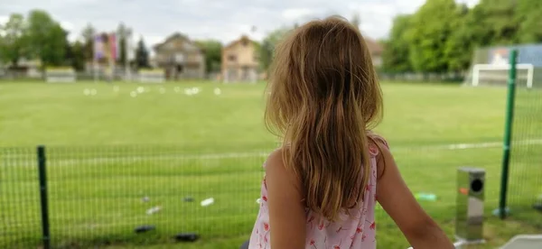 女の子がカメラに背を向けて座って 子供たちのサッカーの試合を見ている 美しい長いブロンドの髪 試合中のサッカーファン — ストック写真