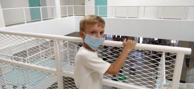 Beyaz gömlekli ve maskeli bir çocuk metal bir çitin yanında duruyor. 7 yaşında bir çocuk tırabzana tutunuyor. Beyaz duvarları olan modern bir okul ya da klinik. Bir çocuk cidden kameraya bakıyor..