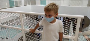 Beyaz gömlekli ve maskeli bir çocuk metal bir çitin yanında duruyor. 7 yaşında bir çocuk tırabzana tutunuyor. Beyaz duvarları olan modern bir okul ya da klinik. Çocuk başka tarafa bakıyor..