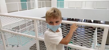 Beyaz gömlekli ve maskeli bir çocuk metal bir çitin yanında duruyor. 7 yaşında bir çocuk tırabzana tutunuyor. Beyaz duvarları olan modern bir okul ya da klinik. Çocuk ciddi ciddi başka tarafa bakıyor..