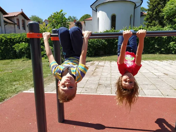 Sremska Mitrovica, Serbia, 20 de mayo de 2020. Los niños juegan y hacen deportes en el patio de recreo. Una chica y un niño de 6 y 7 años cuelgan boca abajo en los bares de gimnasia —  Fotos de Stock