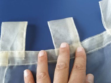 Şeffaf bir organzanın üzerinde bir terzi eli. Tül perdesindeki düğümler. Perde dikiyorum. Mavi arkaplan.