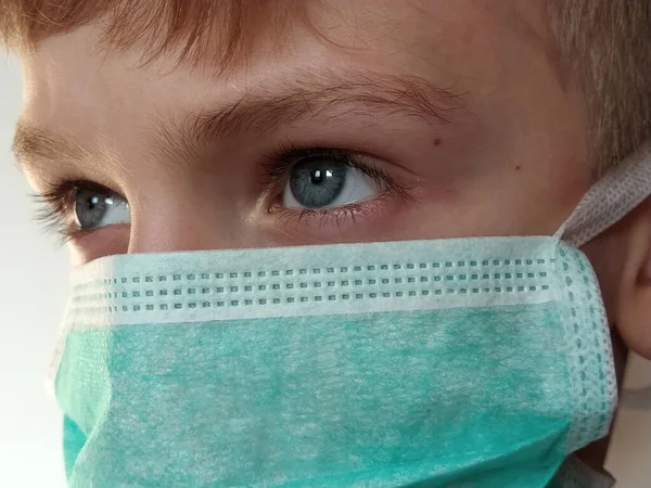 Masked child. The face of a 7-year-old boy in a protective surgical mask close-up. Schoolboy with blond hair and blue-gray eyes on white background.
