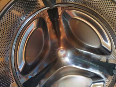 Çamaşır makinesinin varili içeriden. Delikleri ve çıkıntıları olan parlak metal bir tank. Paslanmaz çelikten yapılmış delikli paslanmaz çelik yüzey. Bir çamaşır makinesinin iç görünümü.