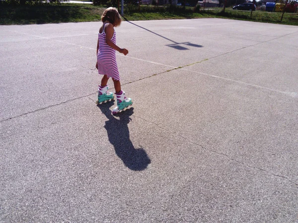 Ta holka má na asfaltu kolečkové brusle. Sedmileté dítě v bílých a růžových pruhovaných šatech jezdí na kolečkových bruslích na hřišti. Letní zábava. Rychlost a trhavé pohyby — Stock fotografie