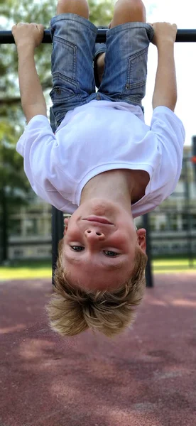 Le garçon est pendu à l'envers sur le terrain de sport. Exercices sur les équipements sportifs. Enfant caucasien de 8 ans aux cheveux blonds. Sport et plaisir — Photo