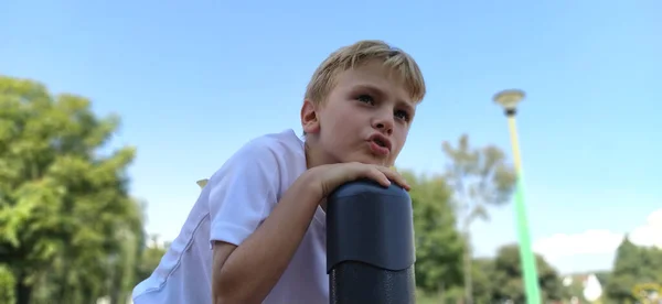 Rapaz pensativo no parque infantil. A criança permaneceu em cima da estrutura metálica, e diz alguma coisa. Menino com cabelo loiro — Fotografia de Stock