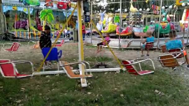 Les enfants heureux montent un carrousel de chaîne dans un parc d'attractions de la ville. Trois enfants se balancent et sourient. Une femme opérateur contrôle la situation et est responsable de la sécurité des enfants. — Video