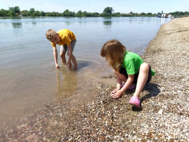 Sremska Mitrovica, Sırbistan, 07 Ağustos 2020. Bir erkek ve bir kız sahilde oynuyorlar. Çocuklar nehir kıyısında su, kum ve çakıl taşlarıyla oynarlar. Çocuklar tişört giyiyorlar. Erkek ve kız kardeş.
