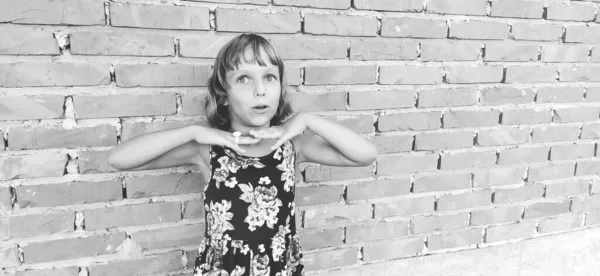 The girl crossed her arms under her chin and laughs. In the background is a brick wall. Caucasian child 7 years old with blond hair. Black and white monochrome photography
