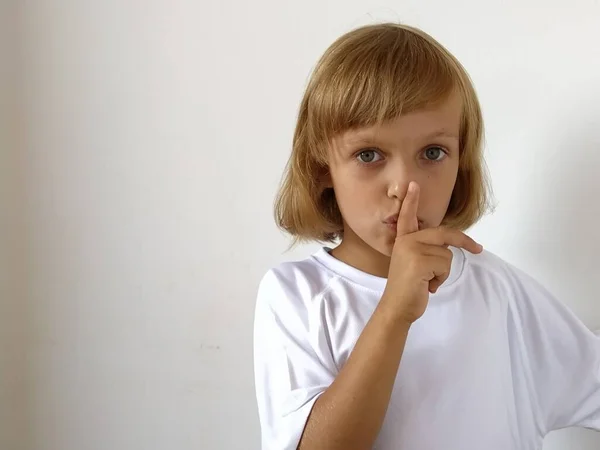 Silence gesture. The finger is near the mouth. The girl on a white background shows a gesture of keeping silence. Child of 7 years old with blond hair. Caucasian type. Copy space. Place for text.
