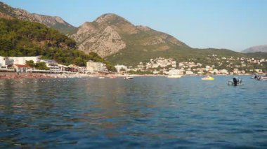 Sutomore, Karadağ, 08.08.2025: Bu video, temiz suların, güneşin tadını çıkaran sahile gidenlerin ve arka planda resimli dağların yer aldığı çarpıcı Adriyatik kıyı şeridini gösteriyor