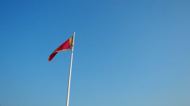 Parlak mavi gökyüzü, canlı renkler ve sembollerle rüzgarda zarif bir şekilde dalgalanan Karadağ bayrağı için bir zemin görevi görüyor. Bu sahne Karadağ 'ın manzara ve kültürünün güzelliğini yansıtıyor.