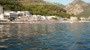 Sutomore, Karadağ, 08.08.2025: Ziyaretçiler Adriyatik kıyı şeridinde dinleniyor, sıcak güneşin, berrak suların ve çarpıcı dağ manzaralarıyla çevrili canlı sahil atmosferinin tadını çıkarıyorlar. Tuzlu suda dalgalanmalar