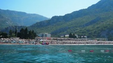 Canj, Karadağ 08.10.2025: Adriyatik Denizi 'nin açık mavi sularının tadını çıkarın. Güneşlenenler kumlu sahilde dinlenirken, nazik dalgalar etrafındaki dağların arasında kumsala doğru süzülüyor..