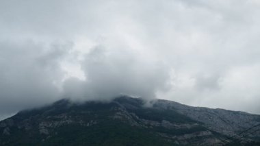 Kara bulutlar dağ sırasını örtüyor. Kalın bulutlar kayalık tepelere yapışıp gizemli bir atmosfer yaratıyor. Yeşillik doğal güzelliğe katkı sağlar. Karadağ, Canj, Bar.