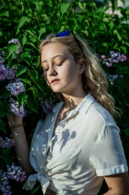 Beyaz gömlekli genç bir kız. Açık, zarif saçlar. Güneş gözlüğü. Çiçek açan leylağın yanındaki parkta duruyor..