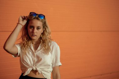 Beyaz gömlekli bir kız turuncu bir duvara karşı poz veriyor. Güneş gözlüklü sarışın. Kızın portresi çok parlak..
