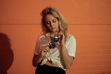 Beyaz gömlekli bir kız turuncu bir duvara karşı poz veriyor. Güneş gözlüklü sarışın. Kızın portresi çok parlak..