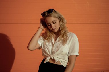 Beyaz gömlekli bir kız turuncu bir duvara karşı poz veriyor. Güneş gözlüklü sarışın. Kızın portresi çok parlak..