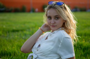 Genç bir sarışın açıklıkta oturuyor. Yeşil çimenler. Bir kız çayırda poz veriyor..