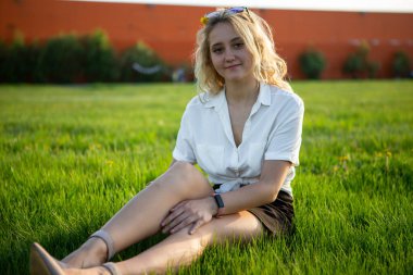 Genç bir sarışın açıklıkta oturuyor. Yeşil çimenler. Bir kız çayırda poz veriyor..