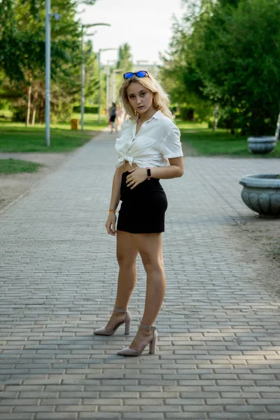 一个穿着黑色裙子和白衬衫的年轻金发姑娘 长长的美丽的腿 站在公园的人行道上 戴著太阳镜 — 图库照片