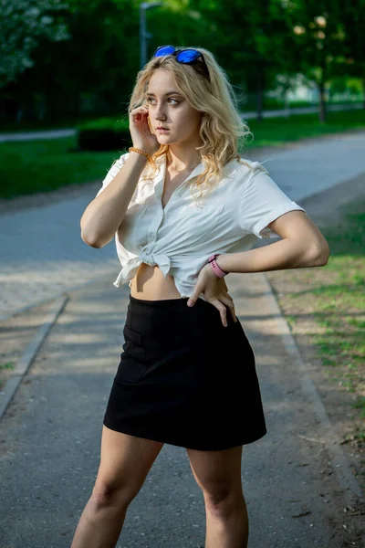 一个穿着黑裙子和白衬衫的少女 轻飘飘的头发太阳镜 它矗立在公园的沥青路面上 — 图库照片