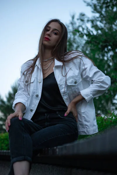 Beyaz ceketli esmer genç bir kız parkta bir bankta oturuyor. Parlak kırmızı ruj çekiciliğe ve çekiciliğe katkı sağlar..