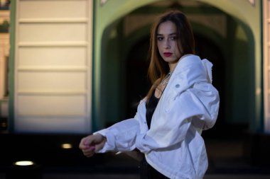 Genç bir kız beyaz ve yeşil bir binanın arka planında poz veriyor. Geceleri esmer bir kadının portresi..