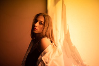 Esmer, gece parlayan sarı bir afişin önünde poz veriyor. Beyaz kot ceketli, siyah tişörtlü, sarı ışıkla aydınlatılmış genç bir kız..