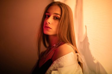 Esmer, gece parlayan sarı bir afişin önünde poz veriyor. Beyaz kot ceketli, siyah tişörtlü, sarı ışıkla aydınlatılmış genç bir kız..