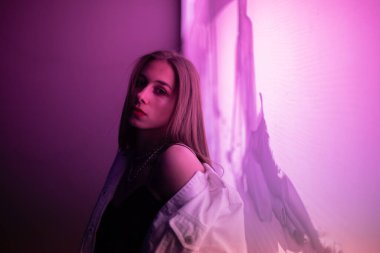 Esmer, gece parıldayan mor bir afişin önünde poz veriyor. Beyaz kot ceketli, siyah tişörtlü, mor ışıkla aydınlatılmış genç bir kız..