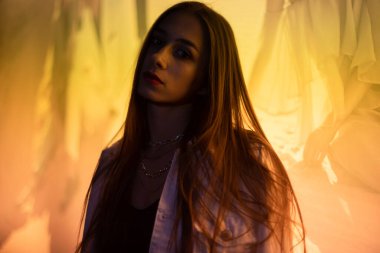 Esmer, gece parlayan sarı bir afişin önünde poz veriyor. Beyaz kot ceketli, siyah tişörtlü, sarı ışıkla aydınlatılmış genç bir kız..