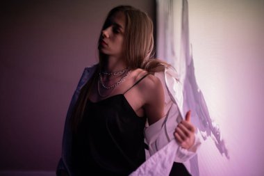 Esmer, gece parıldayan mor bir afişin önünde poz veriyor. Beyaz kot ceketli, siyah tişörtlü, mor ışıkla aydınlatılmış genç bir kız..