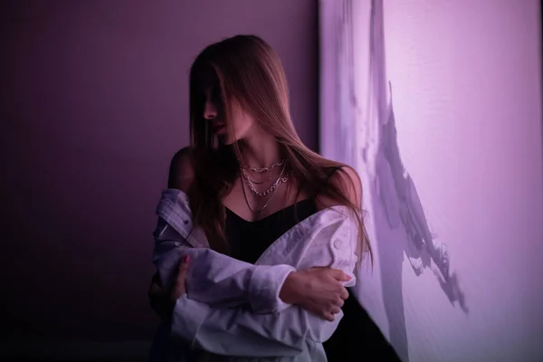 Esmer, gece parıldayan mor bir afişin önünde poz veriyor. Beyaz kot ceketli, siyah tişörtlü, mor ışıkla aydınlatılmış genç bir kız..